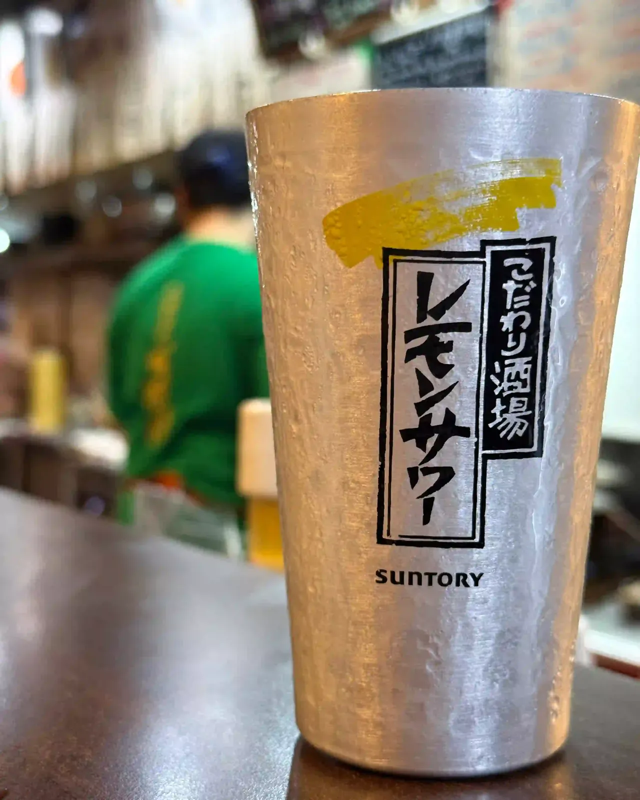 Ein Aluminiumbecher Zitronen-Sour mit japanischer Aufschrift auf der Theke, dahinter der Koch im grünen Shirt.
