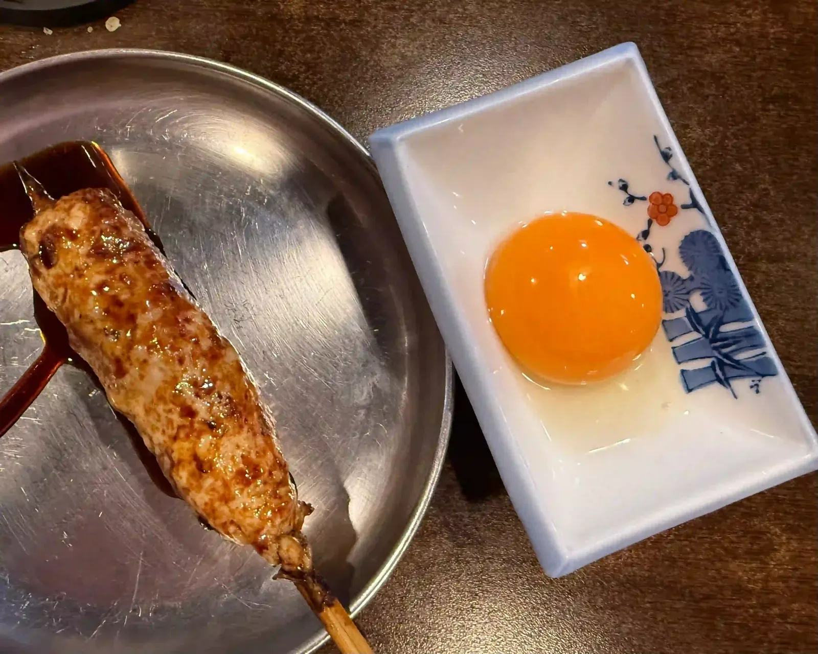 Ein Hackfleischspieß mit Glasur auf einem Blechteller, daneben ein rohes Eigelb in einem Porzellanschälchen.
