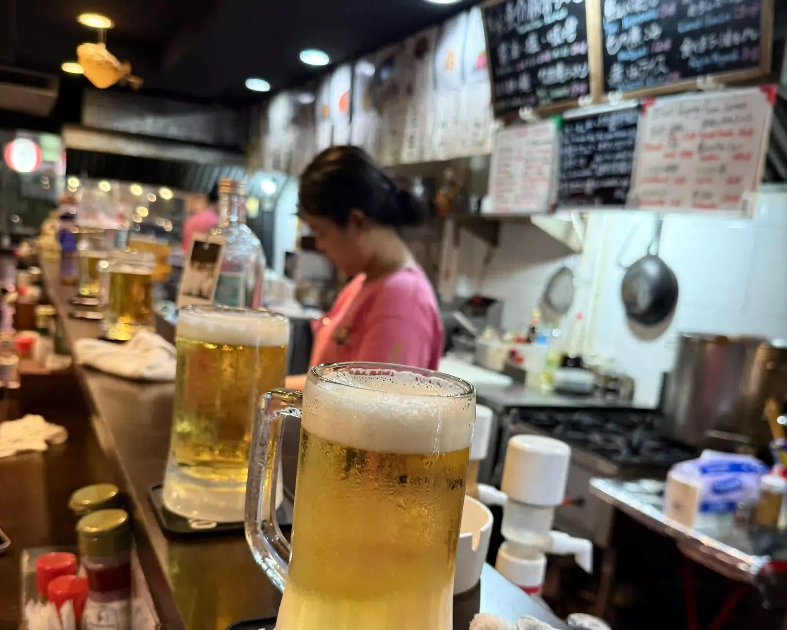 Zwei Bierkrüge auf der Theke, dahinter die Köchin in der offenen Küche unter handgeschriebenen Tafeln.