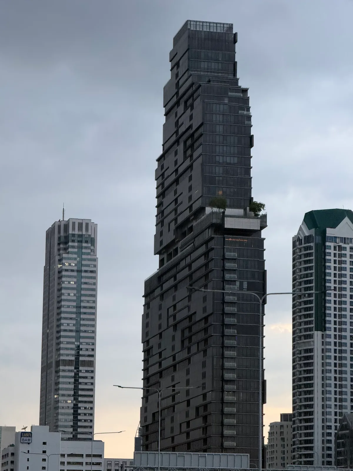 Der Mahanakhon zwischen zwei Hochhäusern vor grauem Himmel.