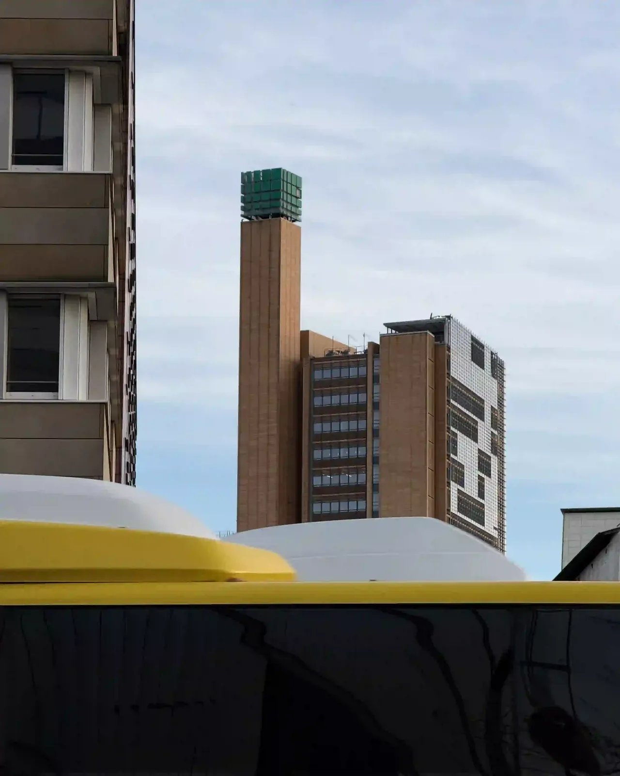 Hinter dem gelben Dach eines Berliner Busses ragt ein Hochhaus mit einem grünen Würfel auf der Spitze in den blassblauen Himmel.